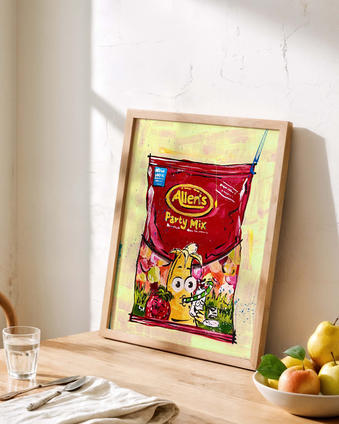 Framed picture of a red bag of Party Mix lollies on a wooden table with a bowl of fruit and glass.