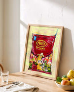 Framed picture of a red bag of Party Mix lollies on a wooden table with a bowl of fruit and glass.